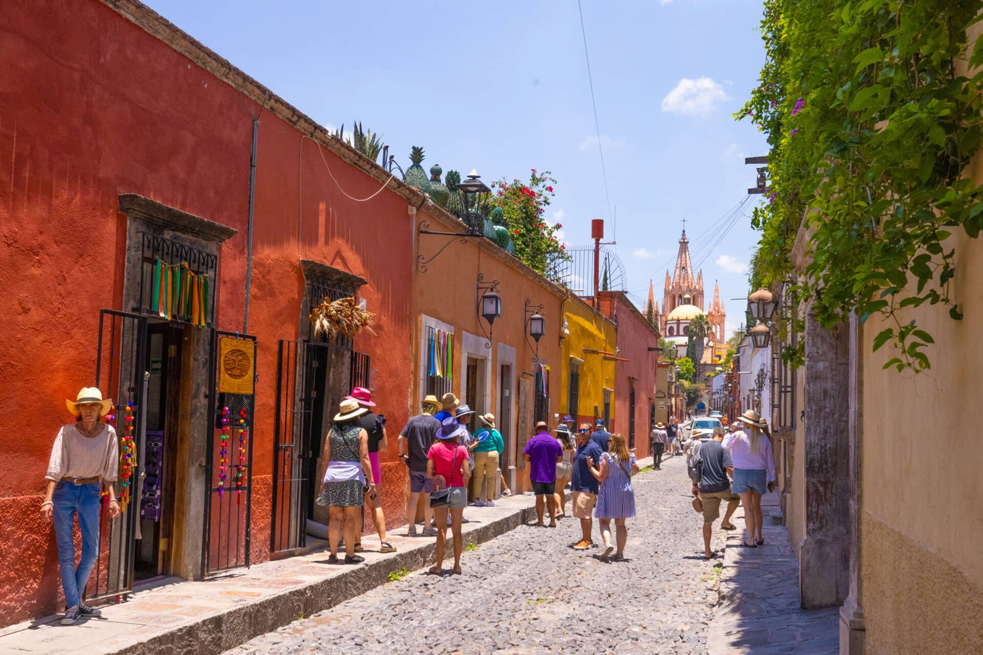 Colorful street in San Miguel de Allende, Mexico, with people walking along cobblestone sidewalks, vibrant buildings, lush greenery, and a church spire—perfect scenery for conference events or relaxing after meetings at nearby hotels and resorts.