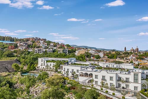 A scenic view of a town with modern white buildings in the foreground, lush greenery, and colorful houses on a hillside—perfect for conference events or relaxing stays at nearby Mexico hotels—under a blue sky with scattered clouds.