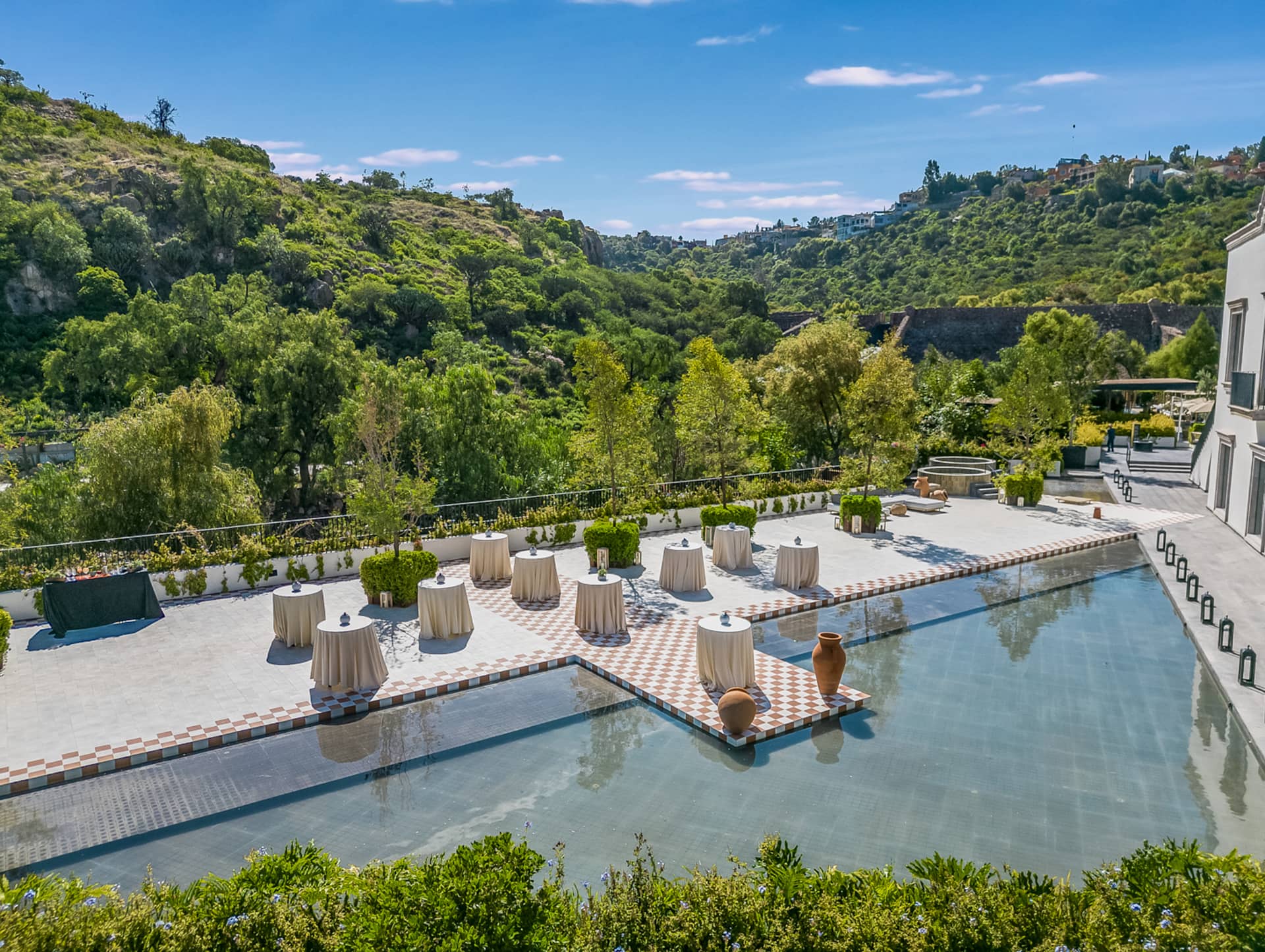 A sunny outdoor event space at a Mexico resort features cocktail tables with beige tablecloths arranged on a patio surrounded by shallow water, perfect for conference events, with green hills and trees in the background.