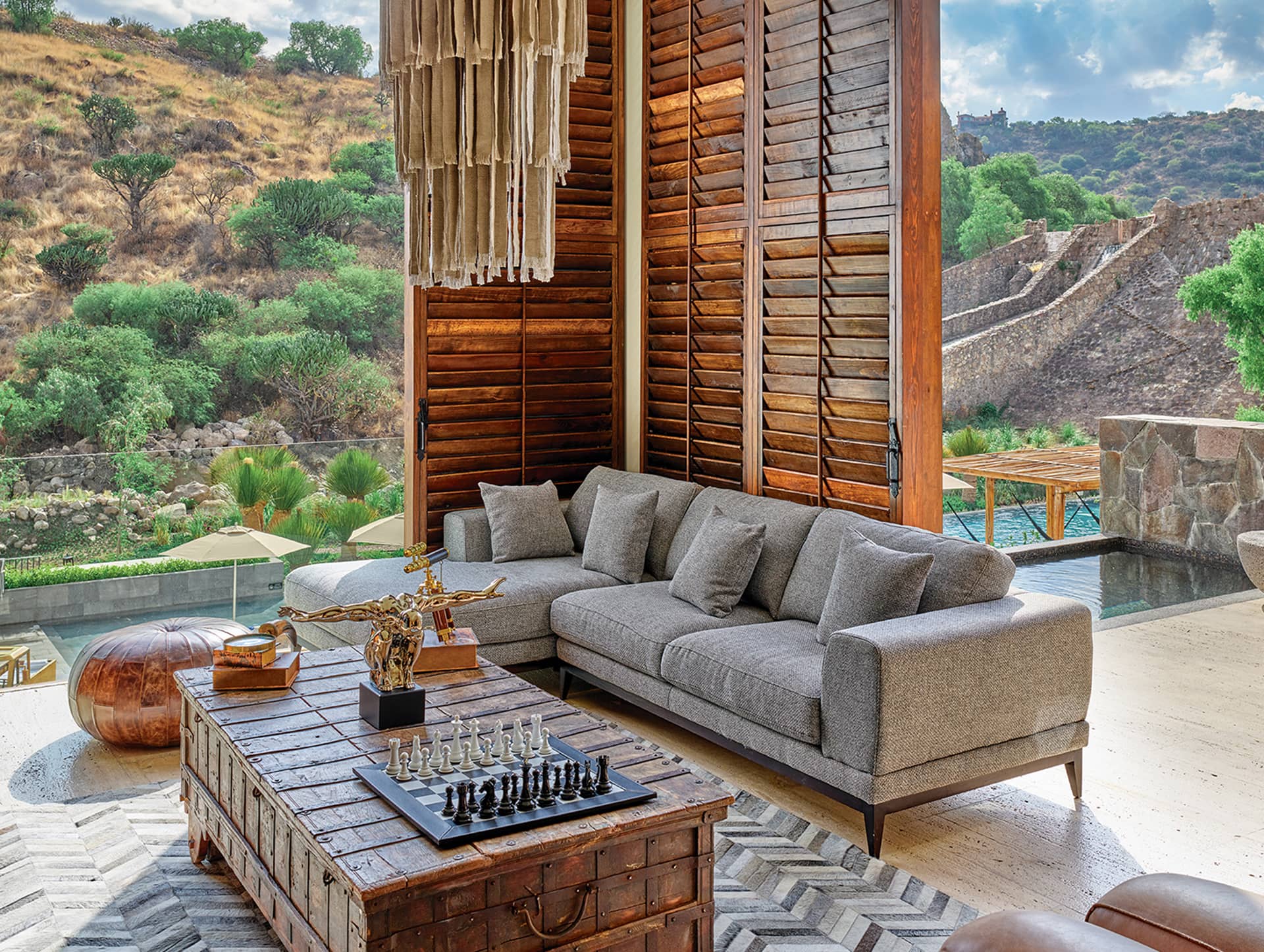 A modern outdoor living space with a gray sectional sofa, wooden coffee table holding a chess set, large wooden shutters, and a pool overlooking a scenic hillside—perfect for informal meetings at top resorts in Mexico.