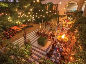 Un patio exterior bellamente iluminado en un complejo turístico de México, con gente cenando en largas mesas, una hoguera central, luces colgantes en forma de estrella, exuberante vegetación y un ambiente festivo y cálido, perfecto para eventos o reuniones al atardecer.