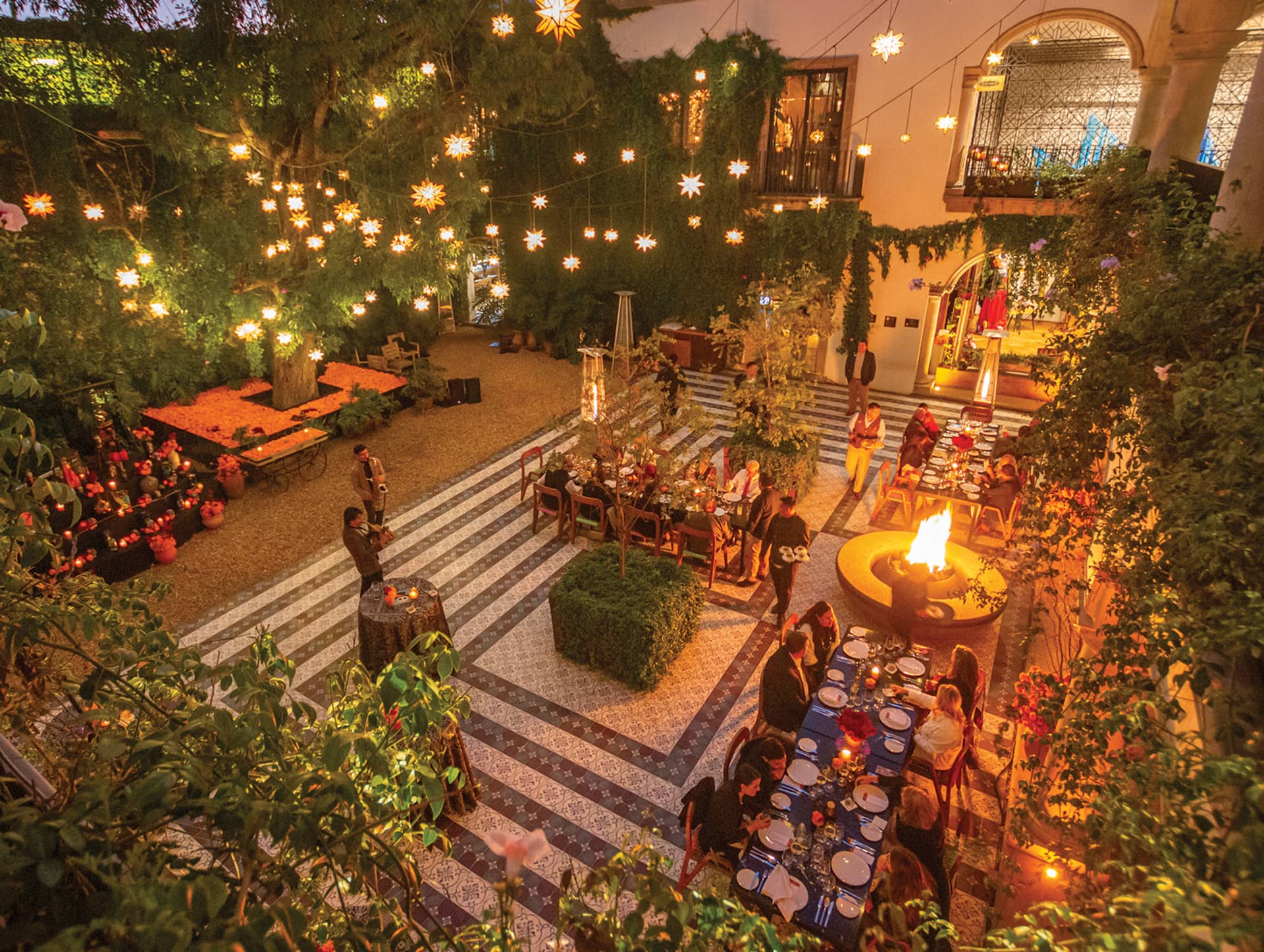 A beautifully lit outdoor courtyard at a Mexico resort, with people dining at long tables, a central fire pit, hanging star-shaped lights, lush greenery, and a festive, warm ambiance perfect for conference events or meetings at dusk.