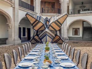 En el patio de uno de los complejos turísticos más elegantes de México, una larga mesa de comedor preparada para una comida formal se alza majestuosamente, con pasillos arqueados y una escultura geométrica de madera como pieza central, perfecta para eventos de conferencias o reuniones de hotel con estilo.