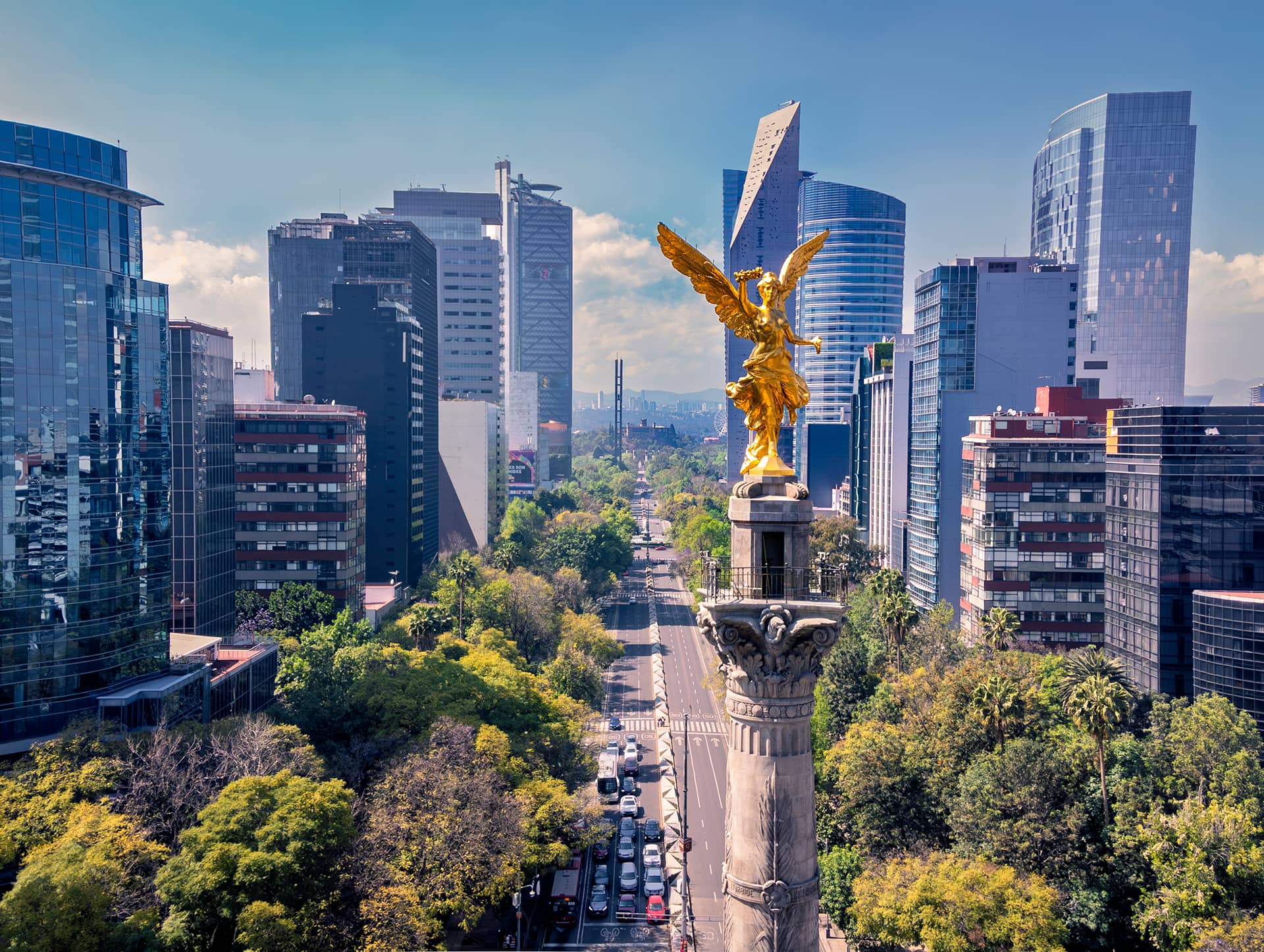 A golden Angel of Independence statue stands atop a tall column, surrounded by modern skyscrapers, lush green trees, and nearby Mexico hotels—an ideal backdrop for conference events and meetings under a clear blue sky.