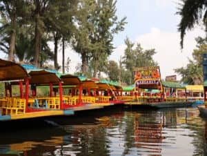 En Xochimilco, México, coloridas barcas con sillas amarillas flotan en un canal rodeado de árboles verdes, cerca de los mejores hoteles y resorts, ideales para conferencias y reuniones. Las barcas están decoradas con colores vivos y una de ellas lleva el nombre de Linda Margarita en su letrero.