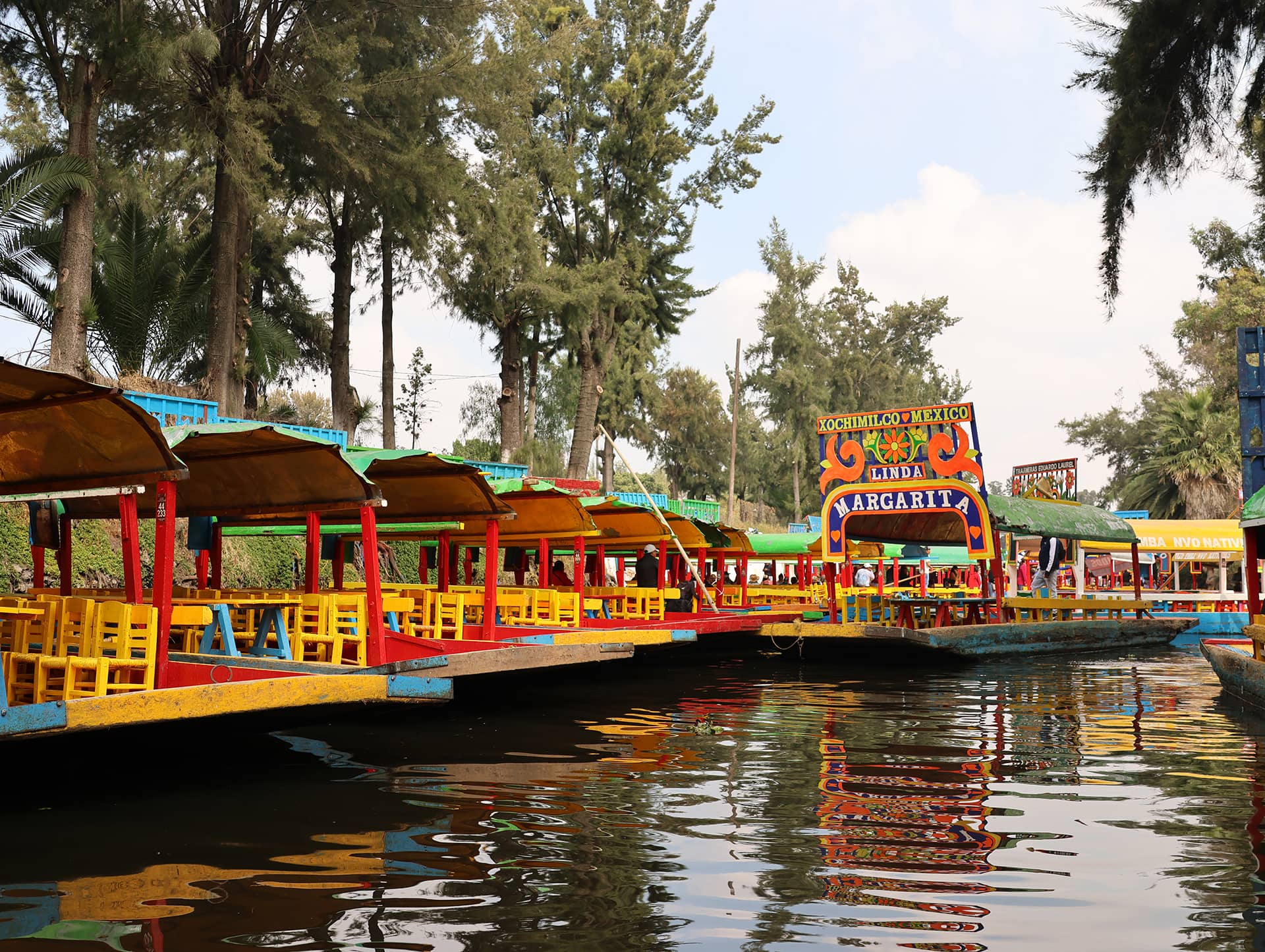 Colorful boats with yellow chairs float on a canal surrounded by green trees in Xochimilco, Mexico, near top resorts and hotels perfect for conference events or meetings. The boats are decorated with bright paint, and one has the name Linda Margarita on its sign.