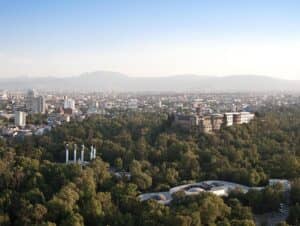 Vista aérea de una gran ciudad con un castillo histórico rodeado de frondosos árboles y parques. Centros de convenciones y hoteles se alzan entre rascacielos, con montañas visibles en el horizonte bajo un cielo despejado.