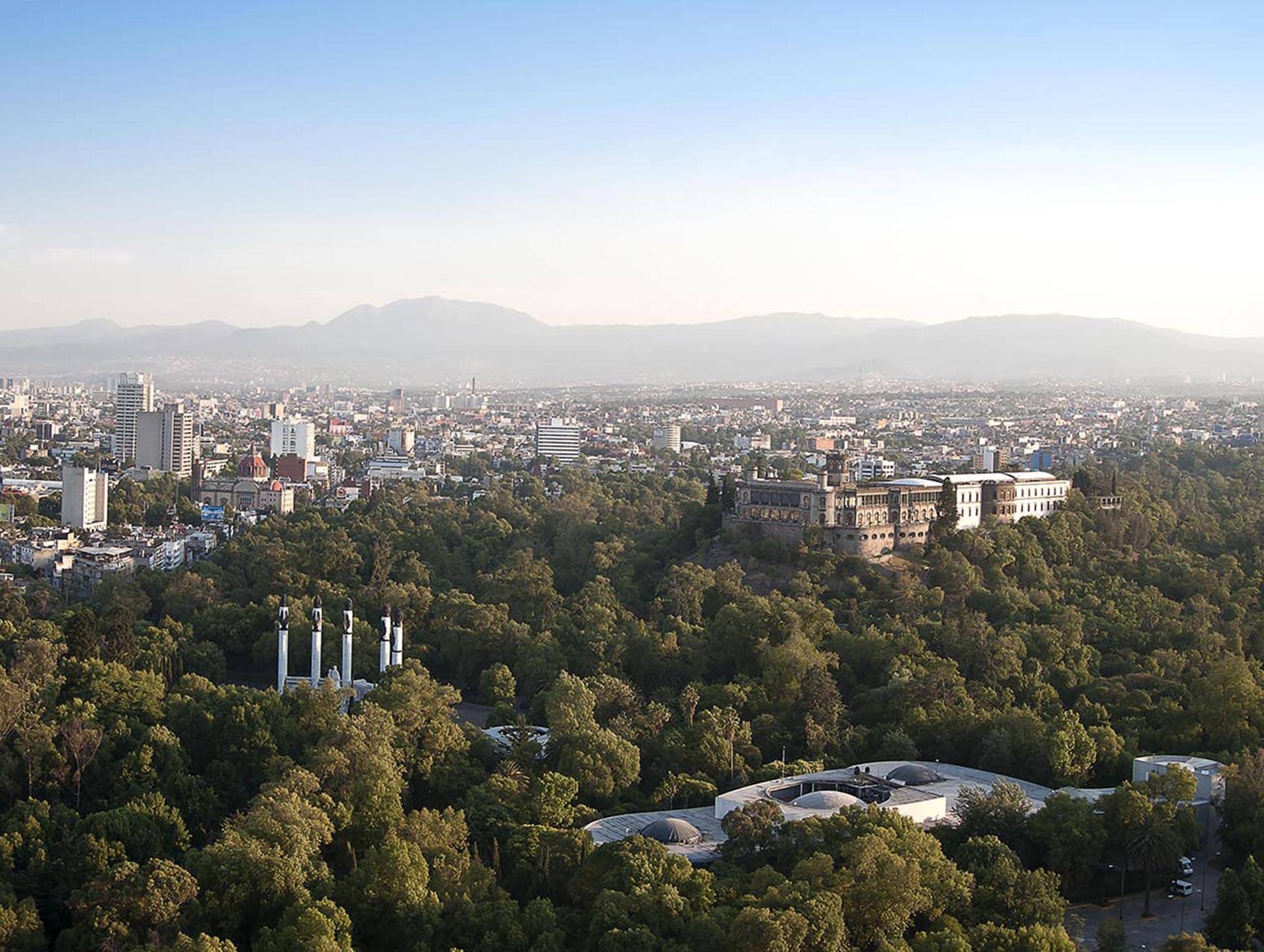 Aerial view of a large city with a historic castle surrounded by dense trees and parks. Conference events venues and hotels rise among skyscrapers, with mountains visible on the horizon under a clear sky.