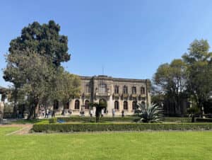 Un histórico edificio de piedra con ventanas arqueadas y balcones se alza tras cuidados jardines y grandes plantas de agave: un emblemático hotel mexicano ideal para reuniones y conferencias. Varias personas pasean cerca de la entrada bajo un cielo azul despejado.