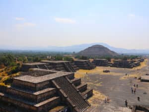 Una vista panorámica de Teotihuacán, que muestra pirámides escalonadas y estructuras de piedra con la Pirámide del Sol al fondo, rodeada de visitantes: un entorno inspirador cerca de hoteles en México, perfecto para reuniones y eventos de conferencias bajo un cielo azul brumoso.