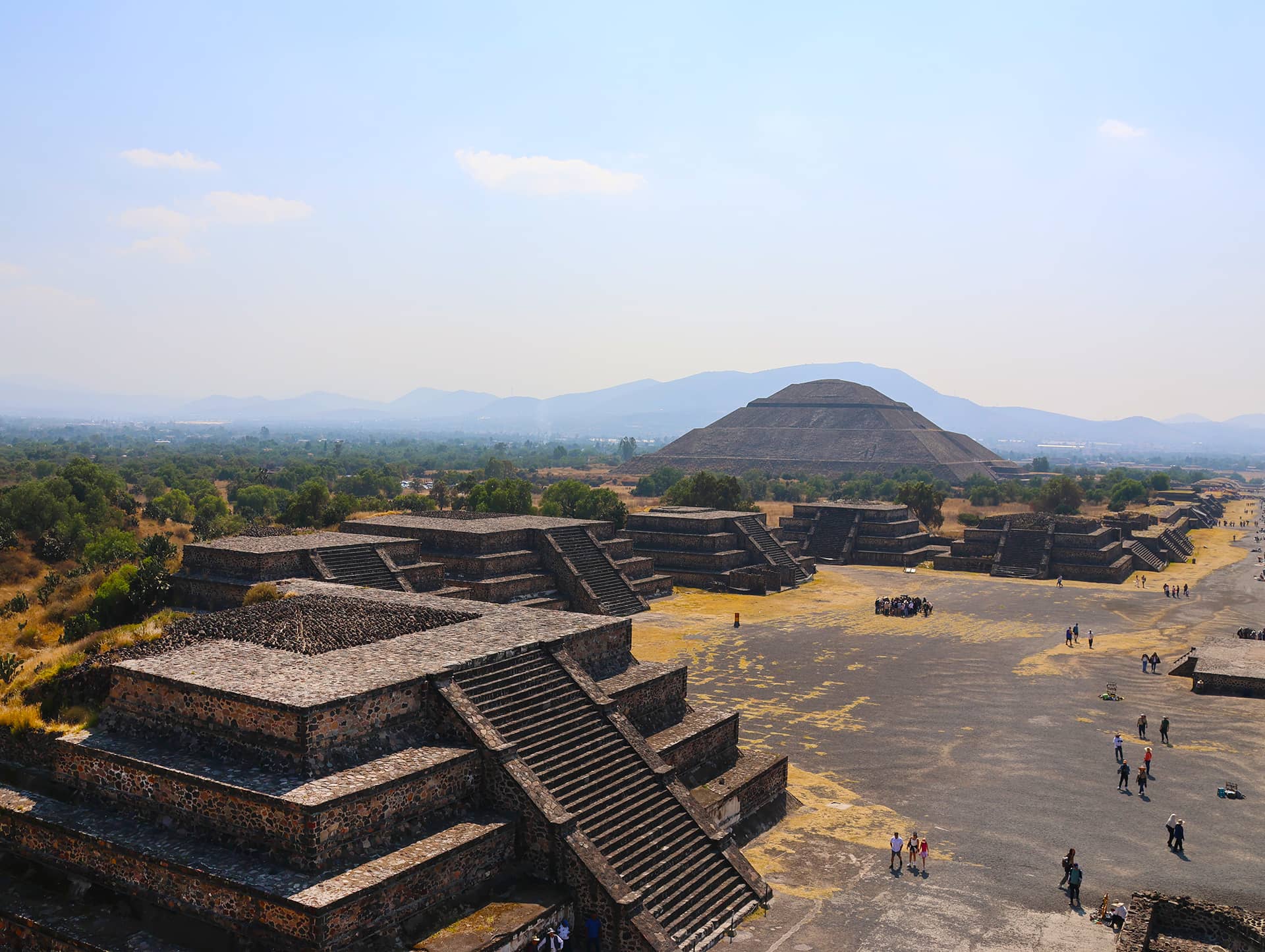 A wide view of Teotihuacan, showing stepped pyramids and stone structures with the Pyramid of the Sun in the background, surrounded by visitors—an inspiring setting near Mexico hotels perfect for meetings and conference events under a hazy blue sky.