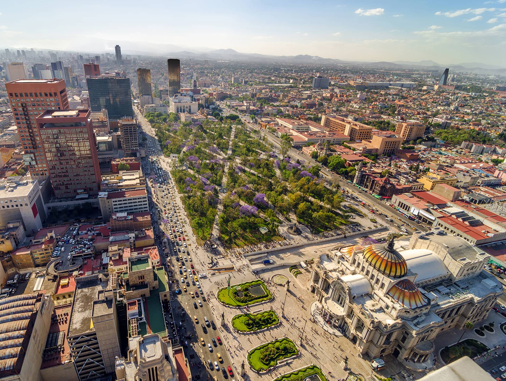 Aerial view of a large city with a wide avenue, green park with purple-flowered trees, and a grand white domed building in the foreground—an ideal setting for conference events or meetings at top Mexico hotels, with distant mountains under a blue sky.