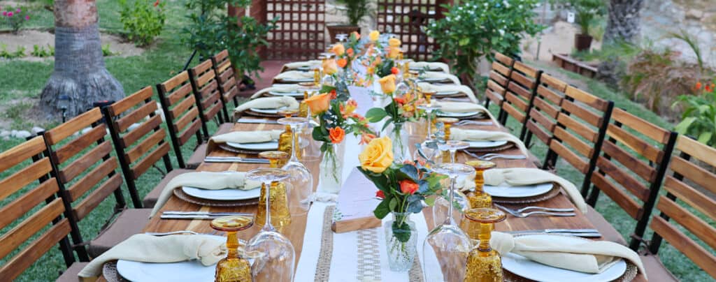 A long outdoor dining table is set with plates, napkins, gold glasses, and vases of yellow and orange flowers. Wooden chairs line both sides, ideal for conference events or meetings at Mexico resorts and hotels in a lush garden setting.
