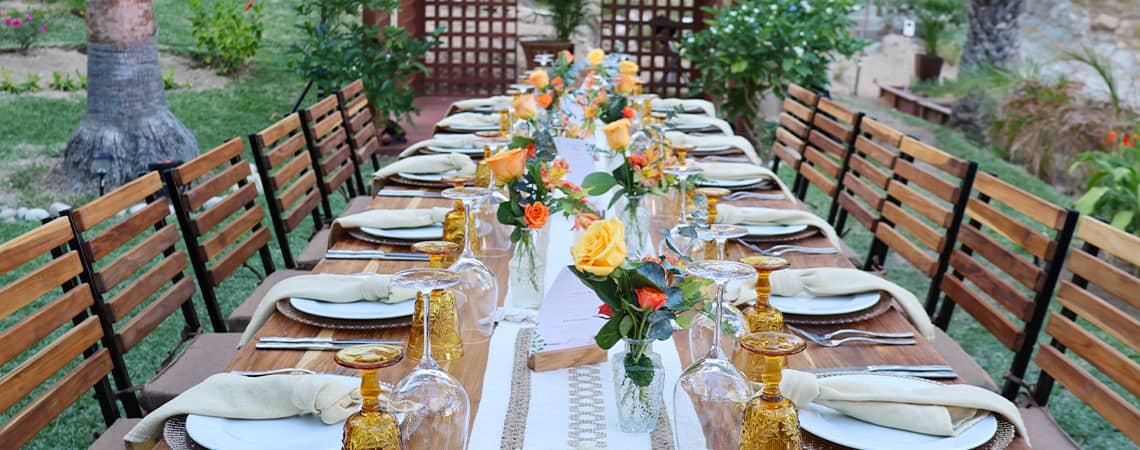 A long outdoor dining table is set with plates, napkins, gold glasses, and vases of yellow and orange flowers. Wooden chairs line both sides, ideal for conference events or meetings at Mexico resorts and hotels in a lush garden setting.