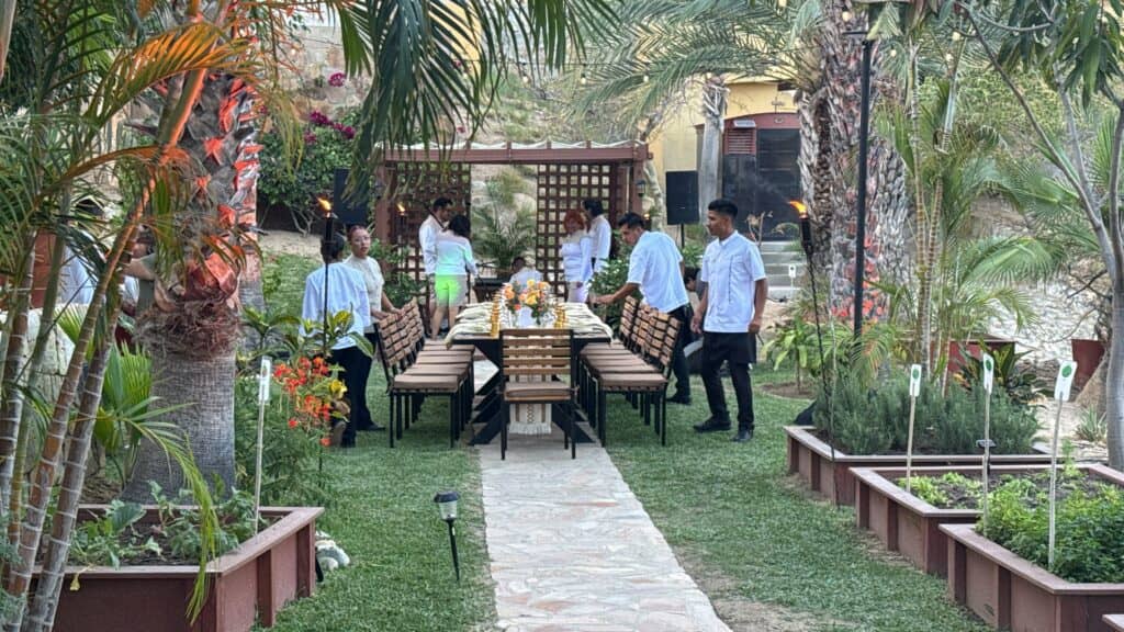 Chefs and servers arrange a long, elegantly set outdoor dining table in a lush garden surrounded by palm trees at one of Mexico’s premier resorts, preparing for an unforgettable conference event.