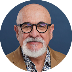 Retrato de un hombre mayor calvo, con barba y bigote blancos, que lleva gafas de montura negra, un blazer color canela y una camisa estampada —perfecto para eventos o reuniones en hoteles de México— sobre un fondo azul liso.