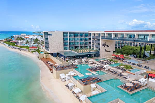 A modern beachfront resort ideal for conferences and meetings, with multiple pools, lounge chairs, cabanas, and palm trees overlooking clear turquoise waters. The building features large windows and a mural of marine animals.