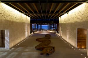 Un amplio y moderno pasillo en uno de los mejores complejos turísticos de México cuenta con paredes de piedra, iluminación empotrada y un banco de madera escultural, que conduce a un área abierta con vistas al océano: un entorno inspirador para conferencias y reuniones.