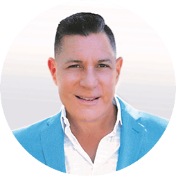 Un hombre de pelo corto y oscuro, que viste una chaqueta azul claro sobre una camisa blanca, sonríe a la cámara con un fondo claro y desenfocado, capturando así el ambiente acogedor de los hoteles mexicanos conocidos por albergar eventos de conferencias memorables.