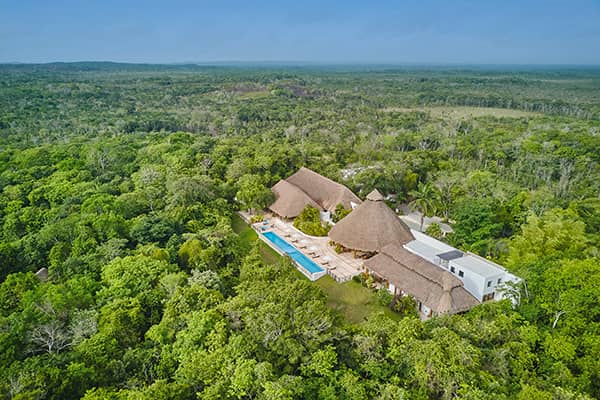 Aerial view of a Conference Events resort with thatched-roof buildings and a long outdoor pool, surrounded by dense green forest under a clear blue sky.