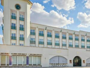 Un gran edificio blanco para conferencias, con múltiples ventanas y balcones, y una alta torre del reloj en el lado izquierdo. El cielo es azul con nubes dispersas.