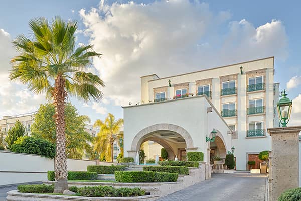 A modern white hotel with green railings, arched entrance, manicured hedges, palm trees, and vintage green lamps—perfect for meetings or conference events—set against a partly cloudy sky.