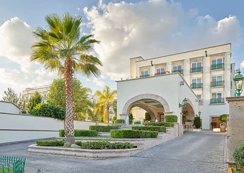 A modern Mexico hotel with white walls and arched entrance, perfect for meetings or conference events, surrounded by neatly trimmed bushes, palm trees, and a paved driveway under a partly cloudy sky.