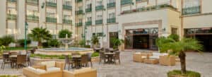 Patio exterior del hotel con asientos de mimbre, mesas y sillas dispuestas alrededor de una fuente central: un espacio acogedor para reuniones en los hoteles de México, rodeado de exuberantes plantas y un edificio de varias plantas con balcones y grandes ventanales.