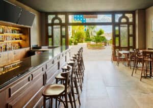 Un moderno bar con mostrador y taburetes de madera pulida se ubica frente a unas puertas ornamentadas que dan a un luminoso patio exterior con vegetación: un lugar perfecto para conferencias o reuniones informales en los mejores hoteles y resorts de México. Las mesas se alinean a lo largo del lado derecho de la sala.