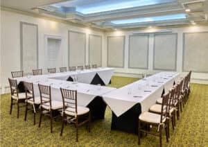Una sala de conferencias en un resort mexicano con mesas en forma de U cubiertas con manteles blancos, ideal para reuniones y eventos. Sillas de madera, jarras de agua, bolígrafos, alfombra estampada y grandes paneles en las paredes completan el ambiente del hotel.