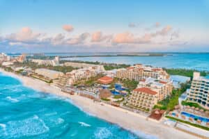 Vista aérea de un complejo turístico frente al mar en Cancún, México, con olas oceánicas de color turquesa, playa de arena blanca, palmeras, varias piscinas y hileras de edificios de hotel, perfecto para eventos y reuniones bajo un cielo azul con nubes dispersas.