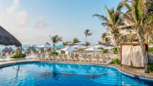 Una piscina de un resort con aguas cristalinas está rodeada de tumbonas, sombrillas y altas palmeras, ideal para eventos y conferencias en hoteles de México. Al fondo, bajo un cielo parcialmente nublado, se divisa el océano y una playa de arena.