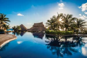 Una piscina tropical en un hotel de México, rodeada de palmeras y cabañas con techos de paja, con el sol brillando intensamente en un cielo azul despejado y su reflejo centelleando en el agua tranquila: perfecta para reuniones o eventos de conferencias.