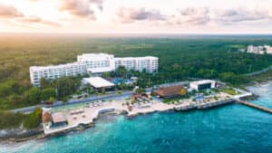 Vista aérea de un complejo turístico frente al mar en México, con un gran edificio de hotel blanco, piscinas y palmeras junto al océano turquesa: un entorno ideal para reuniones o eventos de conferencias en medio de un frondoso bosque y un cielo parcialmente nublado.