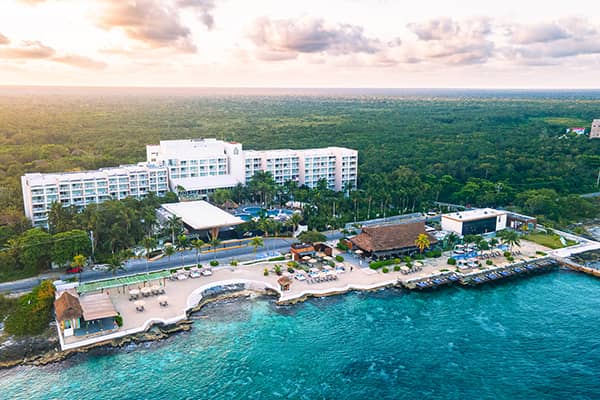 Aerial view of a beachfront conference events resort in Mexico with white buildings, palm trees, pools, lounge areas, and clear turquoise water. Dense green forest stretches out behind the hotel under a partly cloudy sky.