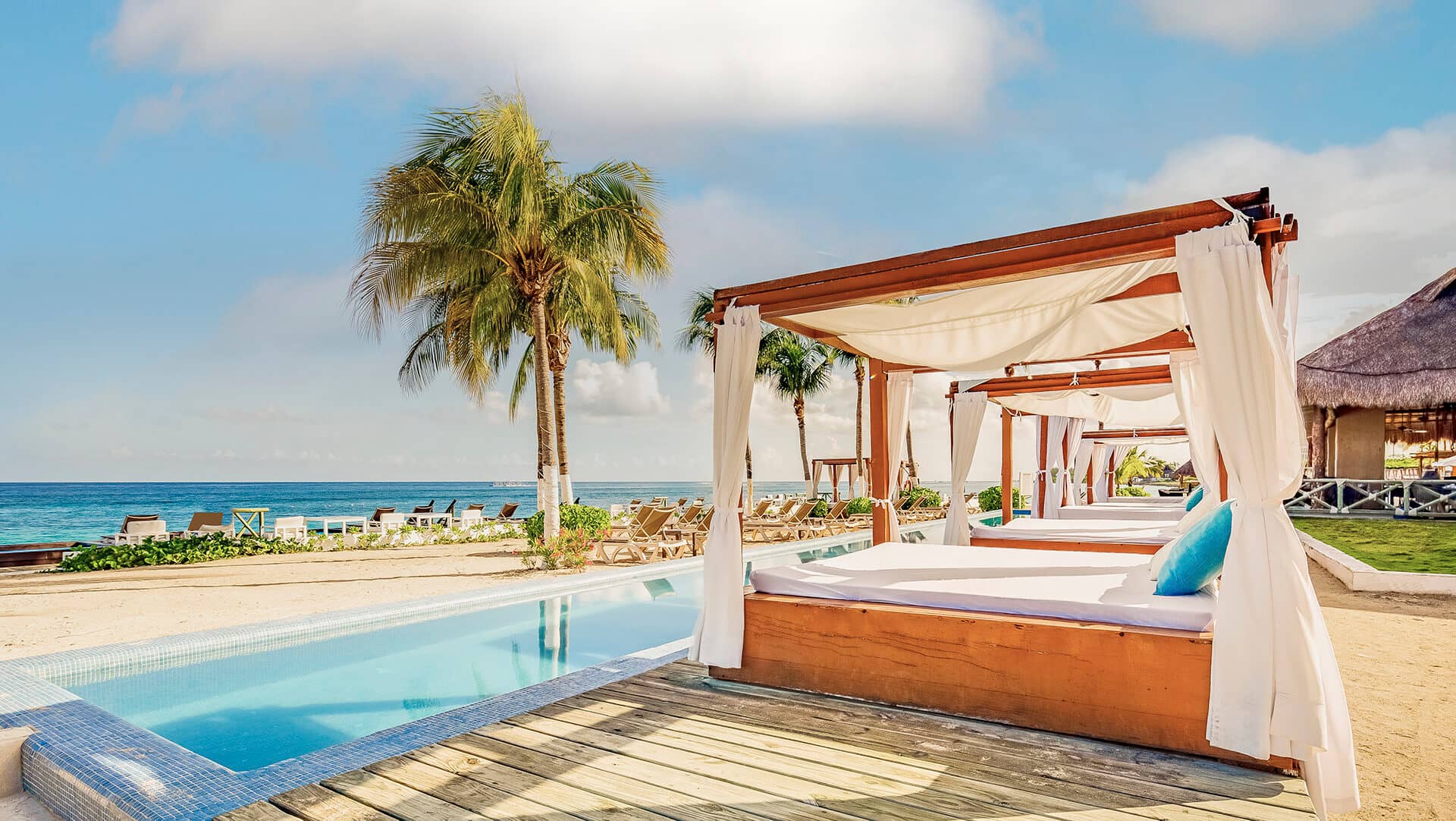 Lujosas cabañas con cortinas blancas bordean una piscina junto a una playa de arena en uno de los mejores complejos turísticos de México, perfecto para reuniones o eventos de conferencias, con palmeras y un océano azul cristalino bajo un cielo brillante de fondo.