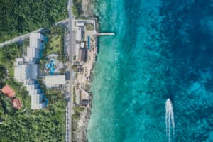 Vista aérea de un complejo turístico costero en México con hoteles rodeados de vegetación, una piscina, un muelle que se adentra en las aguas turquesas del océano y una pequeña embarcación que deja una estela blanca; ideal para conferencias y reuniones.