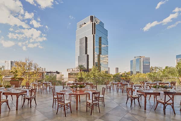 Outdoor terrace with wooden tables and chairs set for dining, surrounded by greenery and city buildings—perfect for conference events or meetings at Mexico hotels. A tall modern glass skyscraper stands out in the background under a blue sky with scattered clouds.