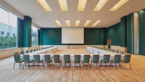 Una sala de conferencias amplia y moderna en los complejos turísticos de México, con paredes verdes, grandes ventanales, una pantalla de proyección y mesas en forma de U rodeadas de sillas verdes: perfecta para reuniones o eventos de conferencias.