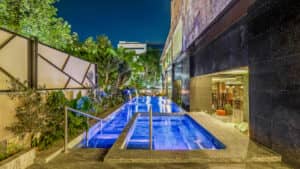 Piscina exterior moderna por la noche, iluminada con luces azules, con jacuzzi incorporado, rodeada de exuberante vegetación y junto a un hotel contemporáneo en México: perfecta para reuniones o eventos de conferencias.