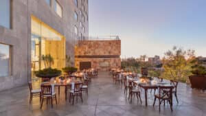 Una elegante terraza al aire libre en un resort mexicano, con mesas redondas preparadas para un evento formal de conferencias, adornadas con velas y sillas cuidadosamente dispuestas. Ubicada junto a un edificio moderno, ofrece vistas de la ciudad y de los árboles al atardecer.