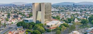 Un alto y moderno edificio de oficinas con la etiqueta Expo Guadalajara se alza sobre un extenso paisaje urbano con numerosas casas, árboles y colinas a lo lejos: un centro ideal para reuniones y eventos de conferencias bajo un cielo despejado.