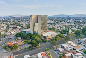 Vista aérea del gran hotel moderno Fiesta Americana, ideal para conferencias y reuniones, rodeado de árboles, calles y edificios más pequeños en una zona urbana con montañas al fondo.