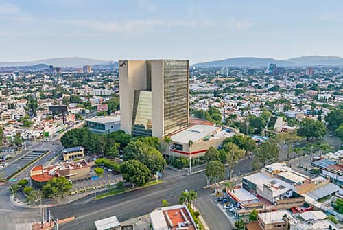 Aerial view of the large modern Fiesta Americana hotel, ideal for conferences and meetings, surrounded by trees, streets, and smaller buildings in an urban area with mountains in the background.