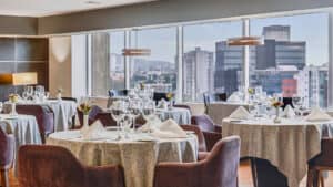 Elegante comedor con mesas redondas preparadas para eventos y conferencias, con manteles blancos, cristalería, servilletas y flores. Grandes ventanales ofrecen vistas panorámicas de modernos hoteles de gran altura. La iluminación tenue crea un ambiente acogedor.