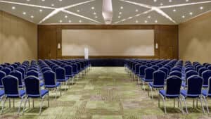 Una sala de conferencias ideal para reuniones en complejos turísticos de México, con filas de sillas azules frente a un escenario y una pantalla en blanco, alfombra verde estampada, paredes beige y un techo con iluminación geométrica y luces en forma de estrella.