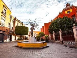 Una estatua de una bailarina indígena con un tocado de plumas se alza sobre un pedestal amarillo en una plaza, rodeada de edificios coloniales y una gran iglesia naranja: una escena inspiradora cerca de los principales centros de convenciones y eventos de México.