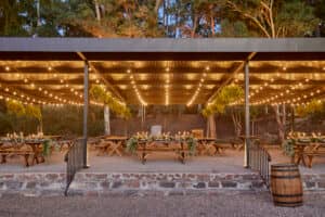 Patio exterior cubierto con guirnaldas de luces, mesas de picnic de madera decoradas con flores y vegetación al fondo. Unas escaleras y barandillas conducen a la zona de estar; a la derecha hay un barril, ideal para eventos y conferencias en los resorts de México.
