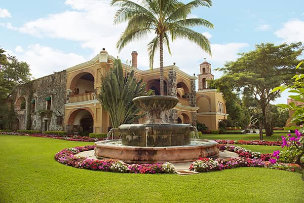 A large stone fountain surrounded by colorful flowers sits on a manicured lawn in front of a historic colonial-style Mexico hotel with arches, balconies, and palm trees under a blue sky.
