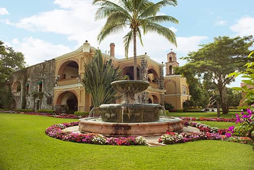 A large historic resort hotel with arched balconies and stone walls stands behind a round fountain, surrounded by palm trees, manicured green lawns, and colorful flower beds—an ideal Mexico setting for meetings or conference events under a partly cloudy sky.